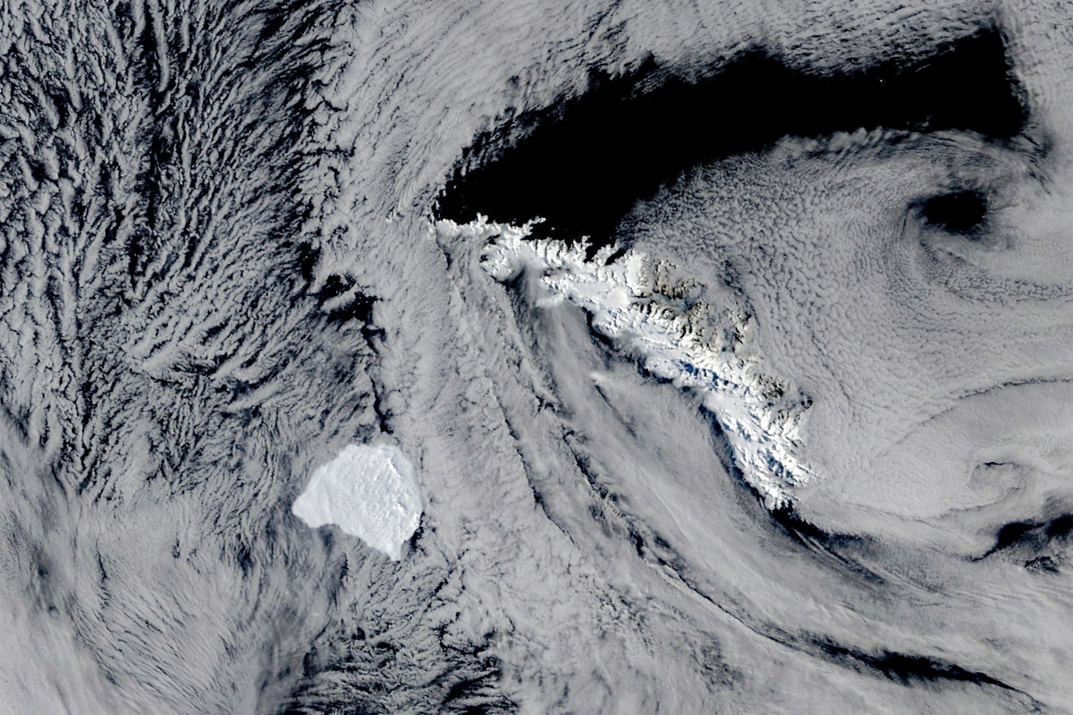 Gran iceberg blanco al suroeste de una isla alargada y nevada, rodeados de océano oscuro y nubes.