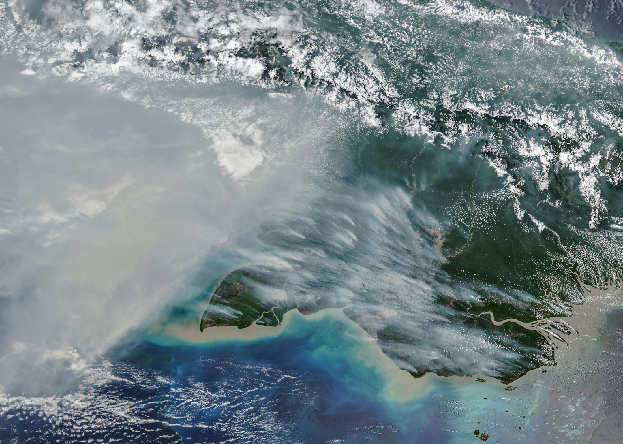Imagem de satélite da costa sul da Nova Guiné, com fumaça cinzenta, rios, mar e nuvens brancas.